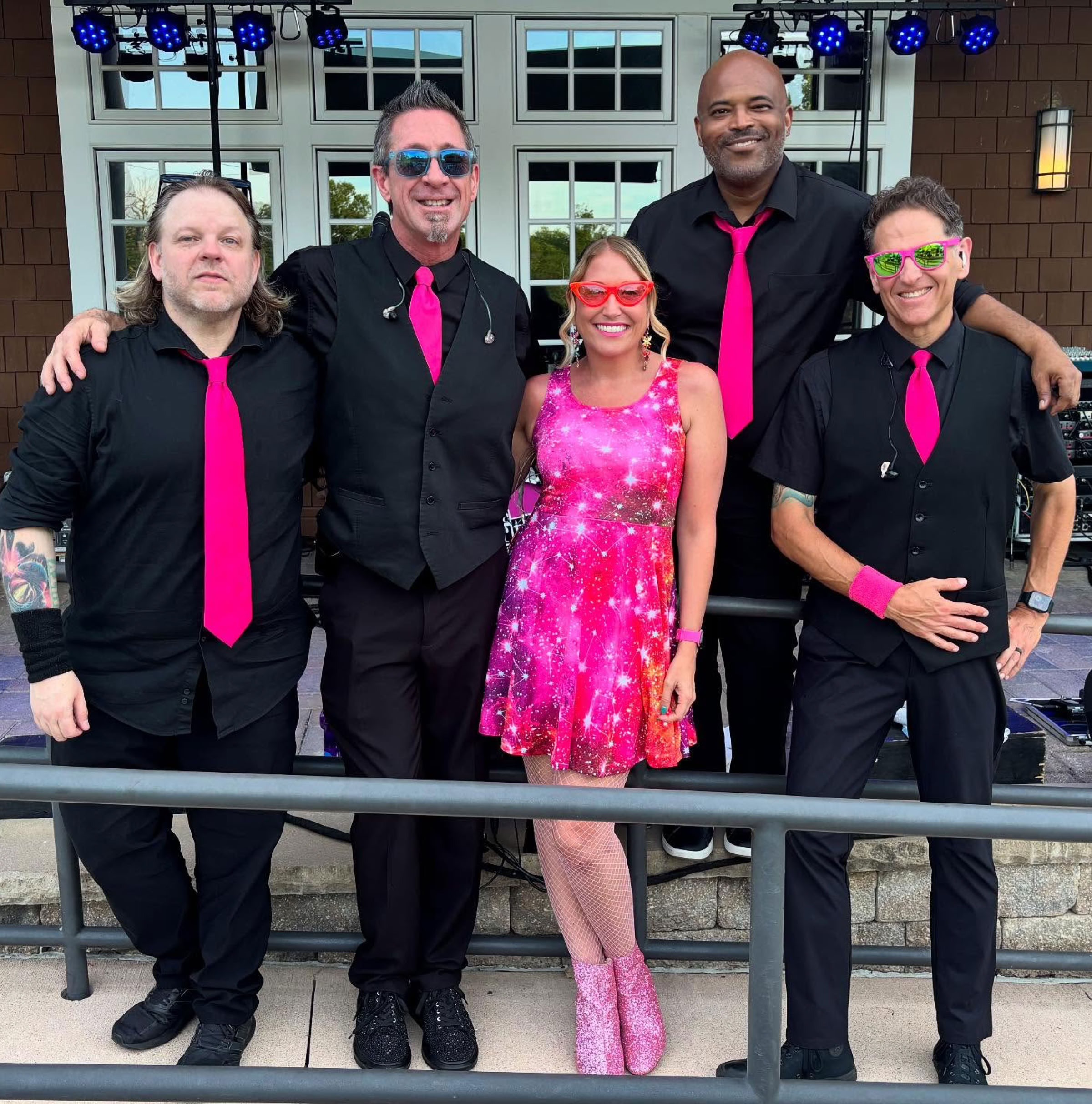 Five people standing in a row, four men wearing black outfits with pink ties and one woman in a pink dress, all smiling in front of a building with windows and stage lights.